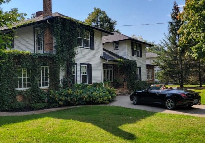 A two-story house with white siding and brick accents, partially covered in ivy, stands near the Jones Big Top. A black convertible is parked in the driveway, while the yard is shaded by green trees under a clear blue sky.