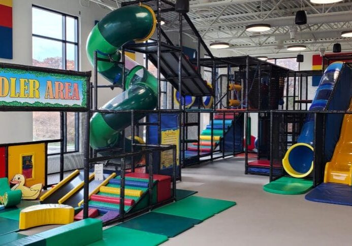 This vibrant indoor playground features colorful slides, climbing structures, and a spiral tube slide. The "Toddler Area" offers soft play equipment and toys for young children in a bright space filled with natural light.