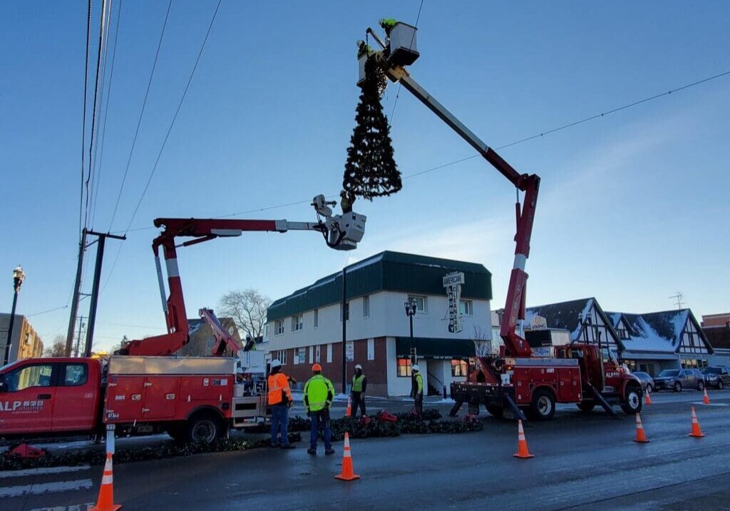 Workers in safety gear use bucket trucks to hang a large Christmas tree decoration above the street for the Lights on Broadway event, with orange cones marking the work area. A two-story building and houses are visible in the background.