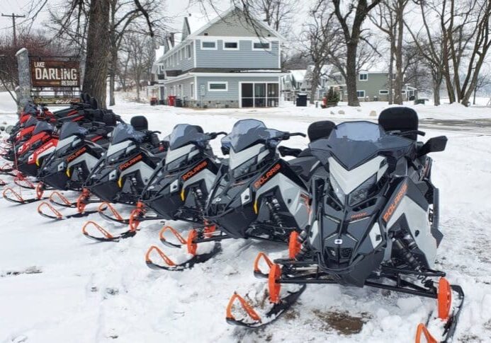 A row of snowmobiles with orange skis is parked on snowy ground in front of houses and bare trees near a sign reading "Lake Darling Resort," showcasing rentals at Lake Darling Resort.