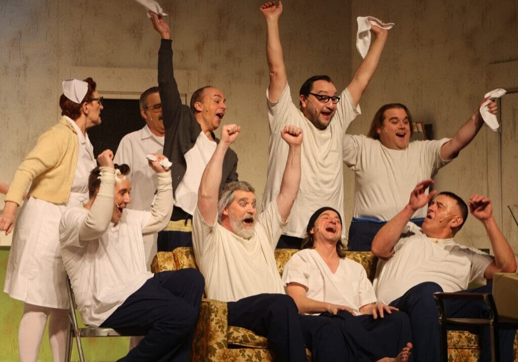A group of people dressed as nurses and patients cheer enthusiastically, raising their arms and waving handkerchiefs, while sitting and standing around a couch in a lively, joyful scene at the Andria Theatre.