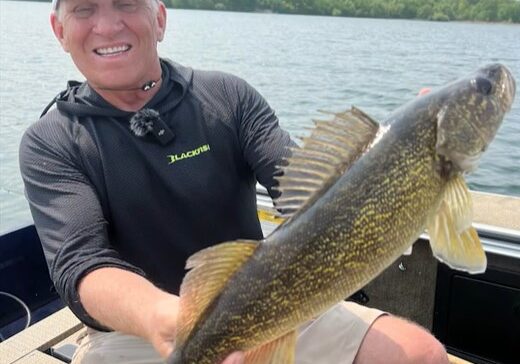 A smiling man in a cap and black shirt sits on a boat, proudly holding a large fish with yellow and brown markings—caught using Multi-Species Swimbaits—with a scenic lake and trees in the background.