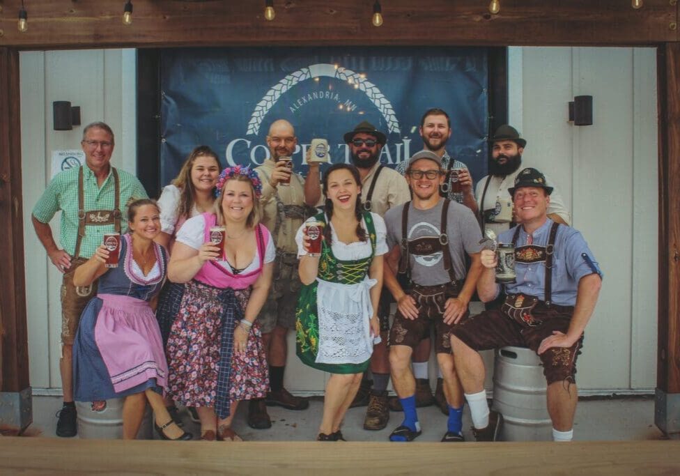 A group of people dressed in traditional Bavarian clothing smile and hold beer glasses, posing together in front of an Oletoberfest banner at a festive event.