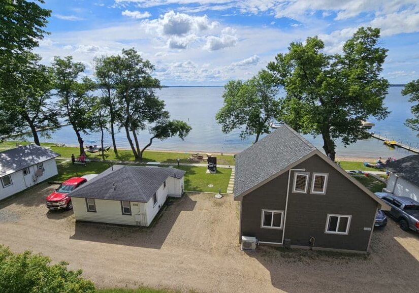 View of lakeside cabins and parked cars with trees and people on a grassy area by the beach at Viking Bay Resort; calm lake water and a dock stretch into the distance under a partly cloudy sky.