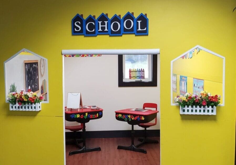 A yellow wall in Grammy's Town is styled like a school entrance, featuring a red clock, "SCHOOL" sign, white fences, flower boxes, and an inviting classroom with desks and colorful decor inside the indoor playground.