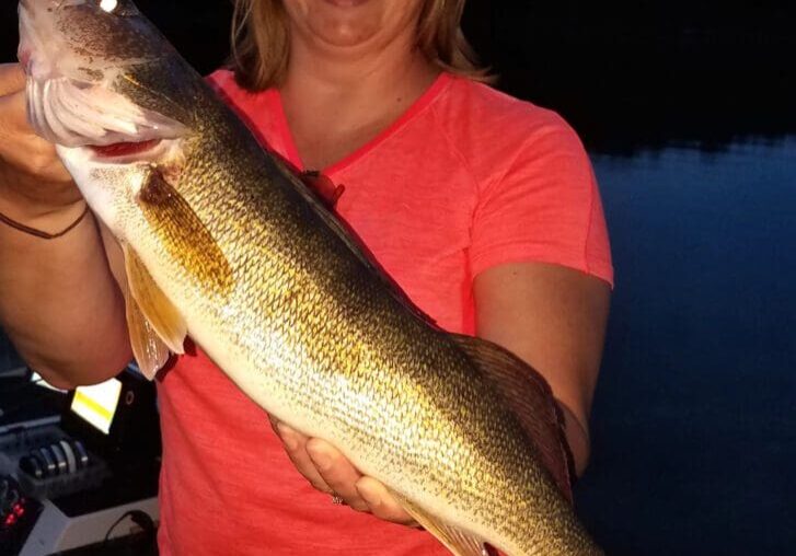 A smiling woman in a pink shirt proudly holds a large fish with both hands while standing on a boat at dusk, framed by dark water and trees in the background.