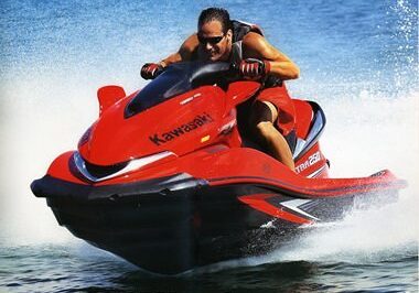 A man wearing sunglasses and a life jacket rides a red Kawasaki jet ski from Fun Rental Express across the water, creating a spray as he speeds forward.