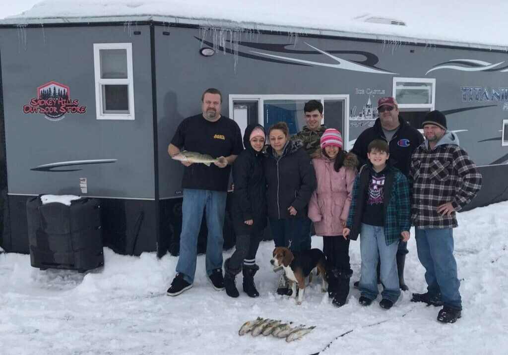 Eight people and a dog pose in front of a dark gray Tyler Wolden Fishing ice fishing trailer on snow, with several fish laid out in front. The group is warmly dressed for winter.
