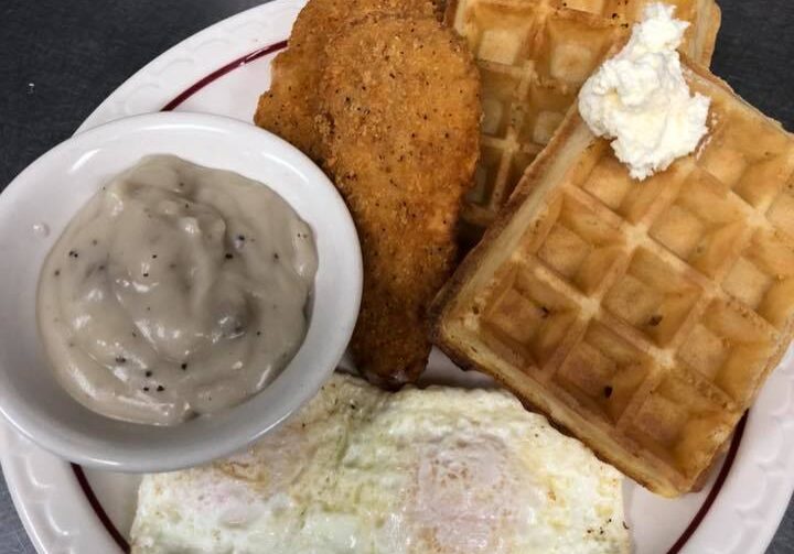 A plate at State Street Cafe featuring two waffles topped with a scoop of butter, two fried eggs, two breaded chicken tenders, and a small bowl of creamy sausage gravy.
