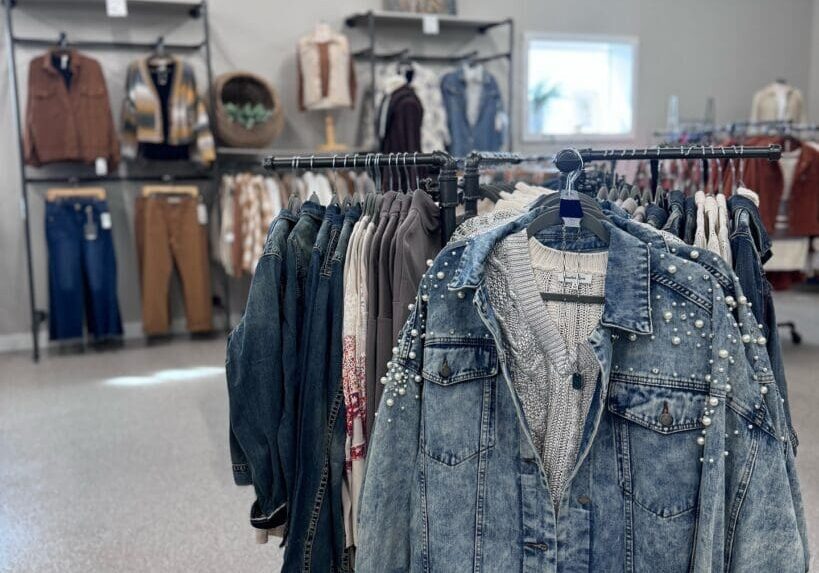 A clothing boutique interior with periwinkle denim and pearl-studded jackets on a rack in the foreground. Racks of shirts, pants, and jackets line the back wall, topped by framed art above.
