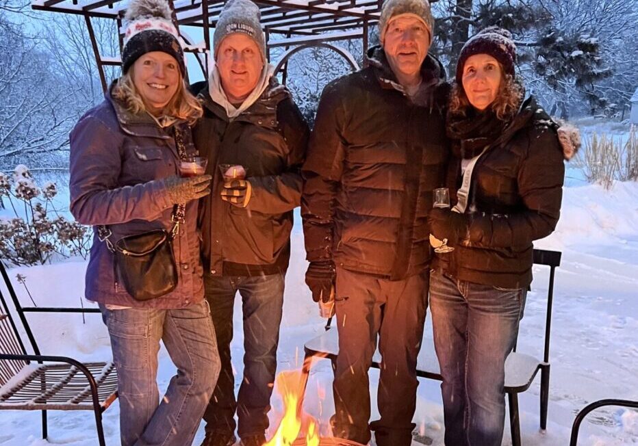 Four adults in winter clothing stand together outside around a fire pit in a snowy backyard, holding drinks and smiling at the camera—capturing the cozy spirit often found at Alexandria Winter Festivals. Snow blankets the ground, trees, and outdoor furniture nearby.