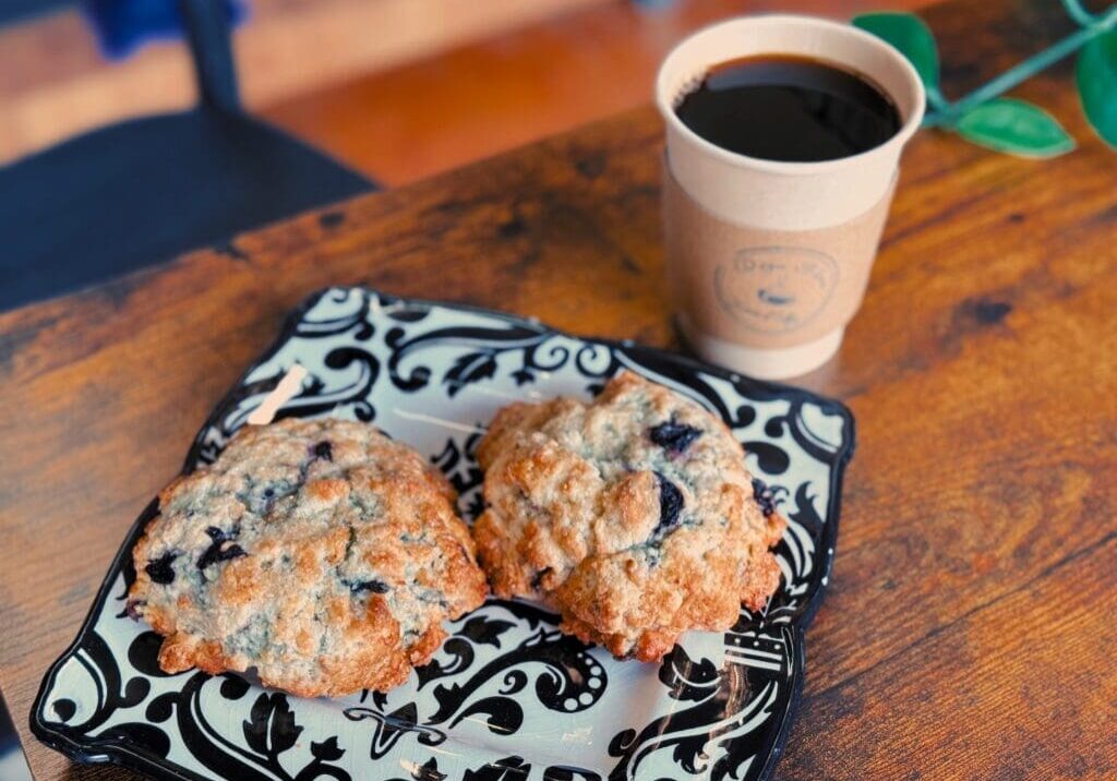 Two blueberry scones on a decorative black and white plate sit beside a cup of black coffee from Deja Bleu Coffee House, all arranged on a wooden table with a green plant partially visible.