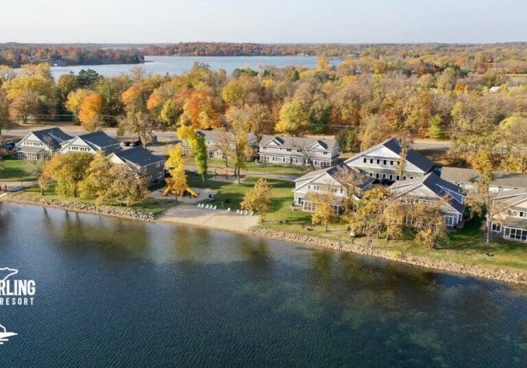 Aerial view of Lake Darling Resort reveals lakeside cabins nestled among autumn trees with vibrant foliage. The calm lake hugs the shoreline, with a dock in view. The "Lake Darling Resort" logo rests in the bottom left corner.