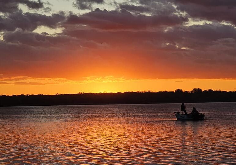 A small boat with two people floats on a calm lake at Eden Acres Resort during a vibrant orange and yellow sunset, with dark clouds overhead and the water reflecting the colorful sky.