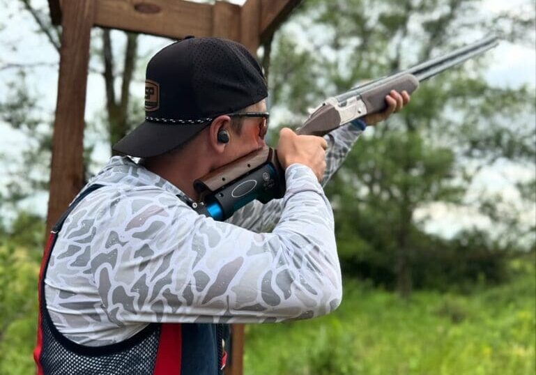 A person wearing a cap, sunglasses, and ear protection aims a shotgun outdoors at Alexandria Shooting Park, standing under a wooden structure surrounded by greenery and trees.