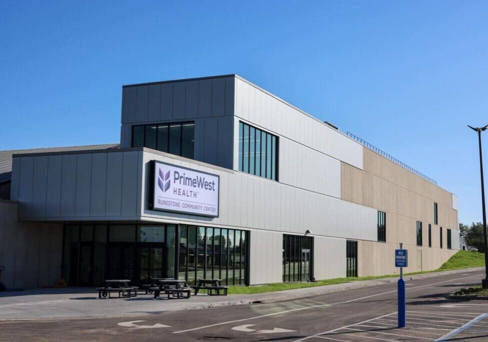 Modern building with large windows and a sign reading "PrimeWest Health, Runestone Community Center" above the entrance; picnic tables sit outside, and the spacious parking lot in front of Runestone Community Center is mostly empty under a clear blue sky.