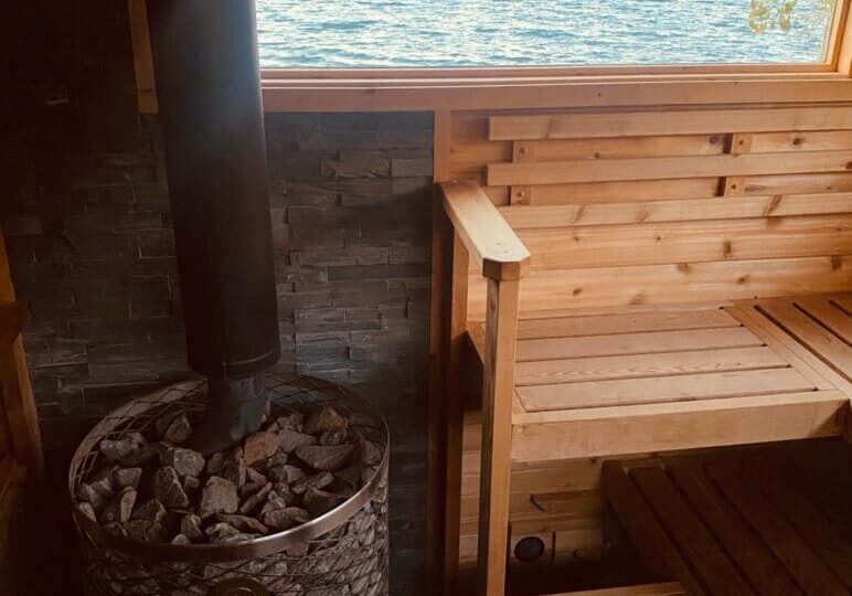 A cozy wood sauna with a stone stove and chimney is pictured indoors. A large window offers a scenic lake view, perfect for relaxing after a Cold Plunge. Wooden benches and walls create a warm, rustic atmosphere reminiscent of Northern Lights Sauna retreats.