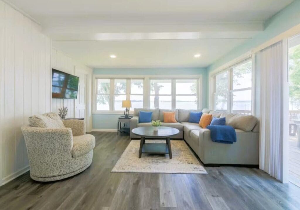 Bright living room at Darling Shores with large windows, an L-shaped sofa with blue and orange pillows, a round coffee table on a rug, swivel chair, wood floors, wall-mounted TV, and gorgeous Lookout views of trees and water.