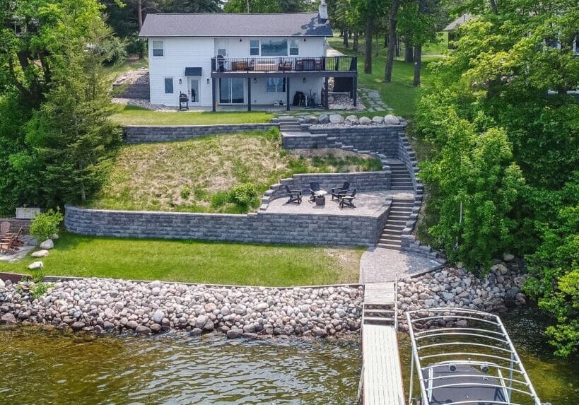 A lakeside house with a large deck overlooks dockside terraced stone landscaping, a fire pit seating area, and a dock with a boat on clear lake water, surrounded by lush green trees and grass.