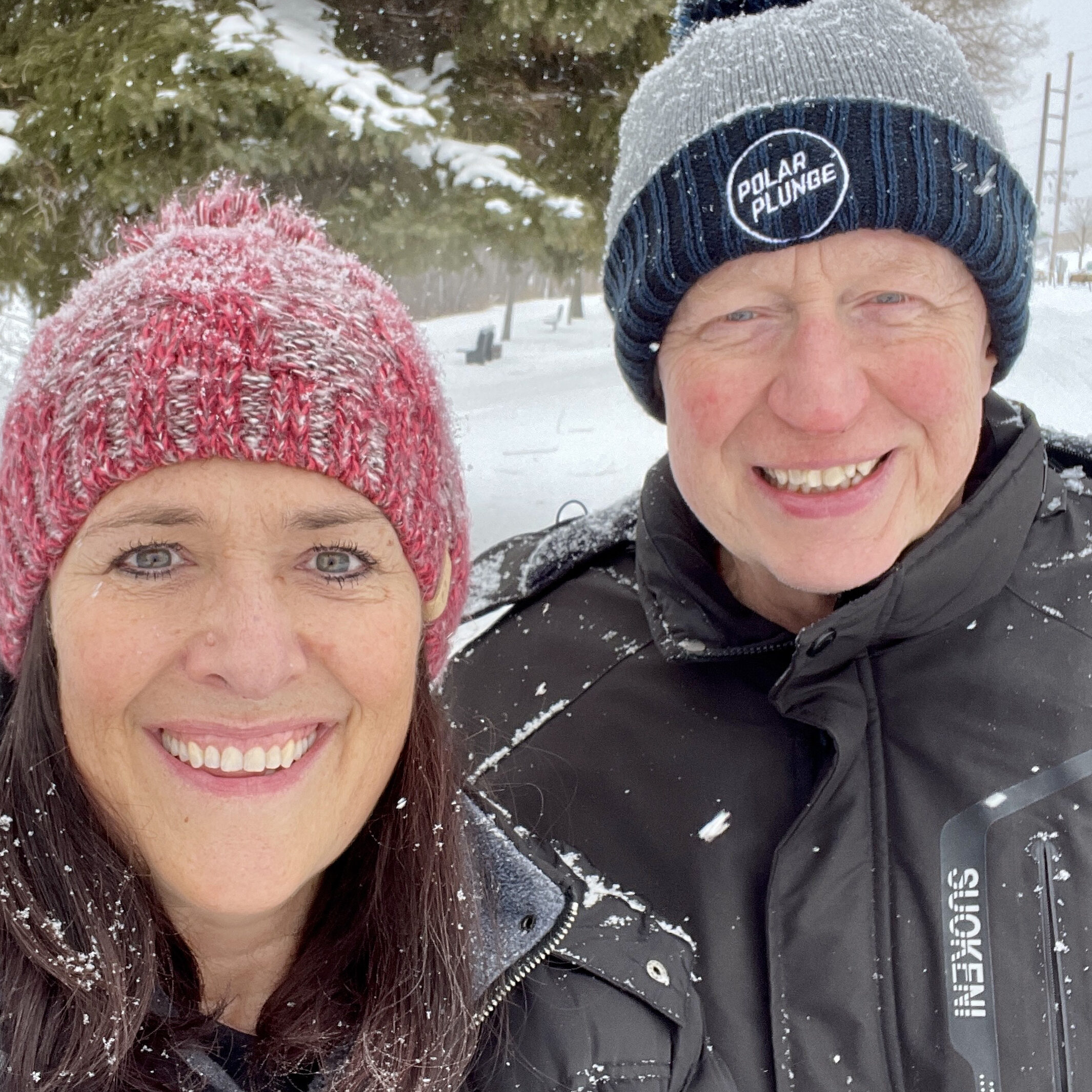 A smiling woman and man in winter hats and jackets stand outside in snowy weather, embodying the spirit of any Winter Guide, with snowflakes on their clothing and trees in the background.