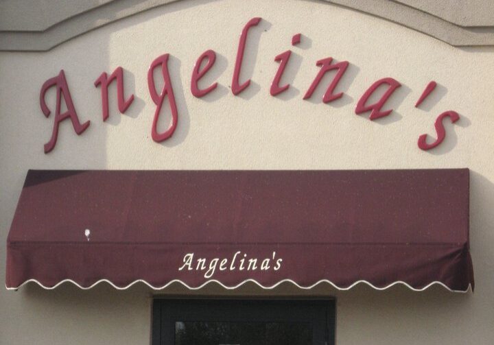 Red script letters spelling "Angelina's" on a beige wall above a maroon awning, which also has "Angelina’s Restaurant" in white script. A black-framed doorway is partially visible beneath the awning, inviting you to enjoy classic Italian food.