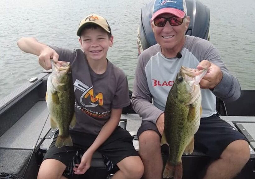 Two people sit in a boat on a lake, smiling and holding up big bass they caught while fishing the Midwest. The younger person wears a cap and T-shirt, while the older one sports sunglasses, a cap, and a long-sleeve shirt.