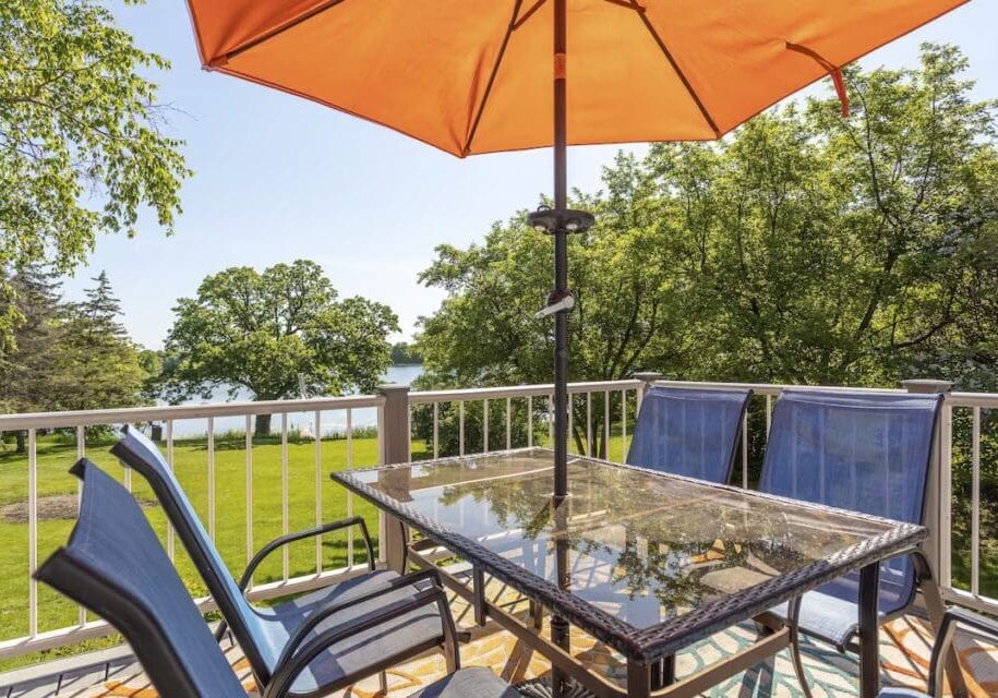 A glass patio table with four blue chairs sits under an orange umbrella on a balcony, perfect for a Taylor-Made Stay, overlooking green trees and a lake in the background on a sunny day.