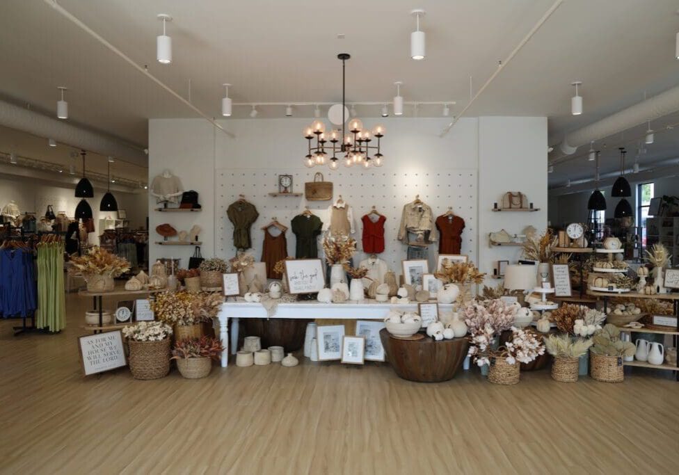 A boutique shop interior at Creative Touch Boutique features dried floral arrangements, baskets, neutral-toned home décor, and framed art on tables and shelves, with clothing and accessories elegantly hung along the back wall.