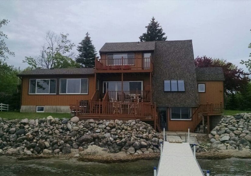 An A-Frame on Lake Darling with large windows and multiple balconies sits on a rocky shoreline. A white dock extends from the house over the water, surrounded by lush trees and greenery.