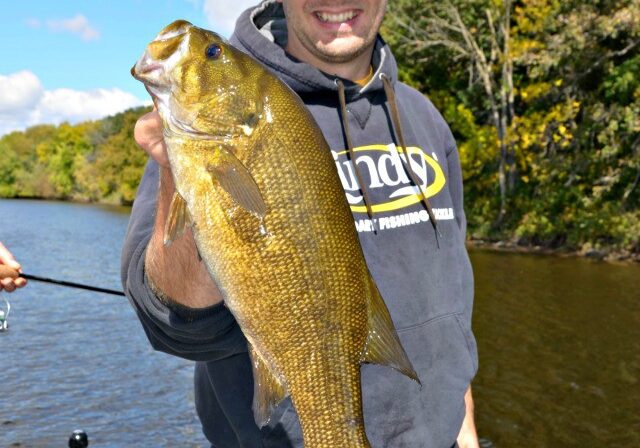 A smiling person in a hoodie and cap proudly holds up a large fish on a boat, enjoying a sunny fishing trip with a river and trees in the background.