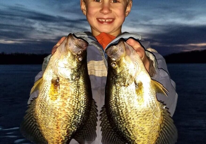 A smiling young boy stands outdoors at dusk, holding up two large, shiny fish caught during an aggressive bite, one in each hand. The sky behind him is darkening with streaks of clouds.
