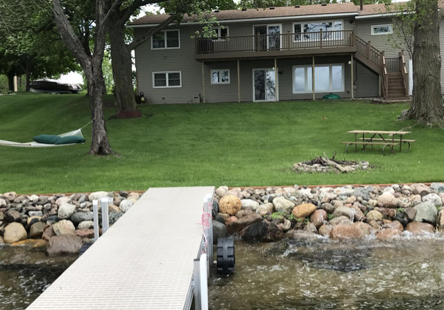 A lakefront AA Cowdry Lake Home with a large grassy yard, hammock, picnic table, and fire pit. A wooden deck and stairs lead to the house, while a dock extends into the lake. Trees surround this peaceful property.