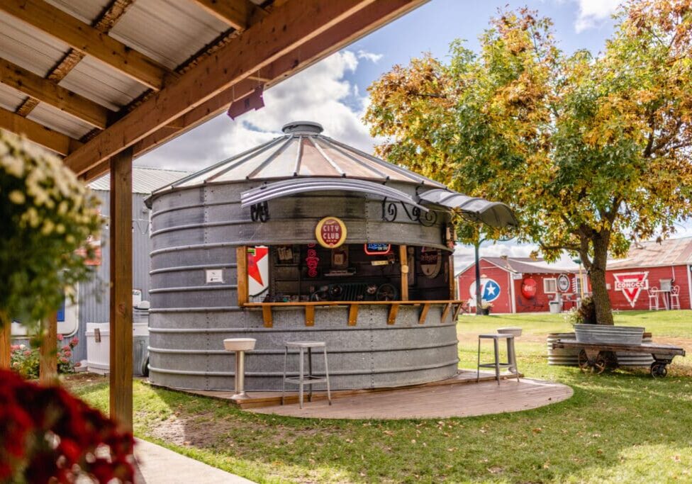 A round metal grain silo is converted into an outdoor bar with stools in front, set in a grassy area at a retro market, surrounded by trees and red barns under a partly cloudy sky.