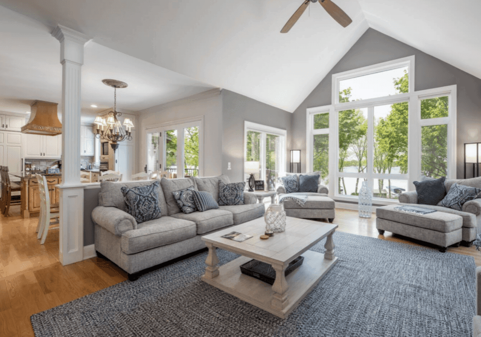 Spacious living room with gray sofas, a light wood coffee table, and large windows revealing Lake Le Homme Dieu—a true lakefront escape—open to a bright kitchen with white cabinets, wood range hood, and light hardwood floors throughout.
