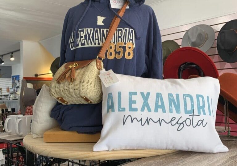 A display featuring a blue "Alexandria 1858" hoodie, a straw handbag, and a white "Alexandria Minnesota" pillow inside Art & Soul, with hats, mugs, and other accessories in the background.