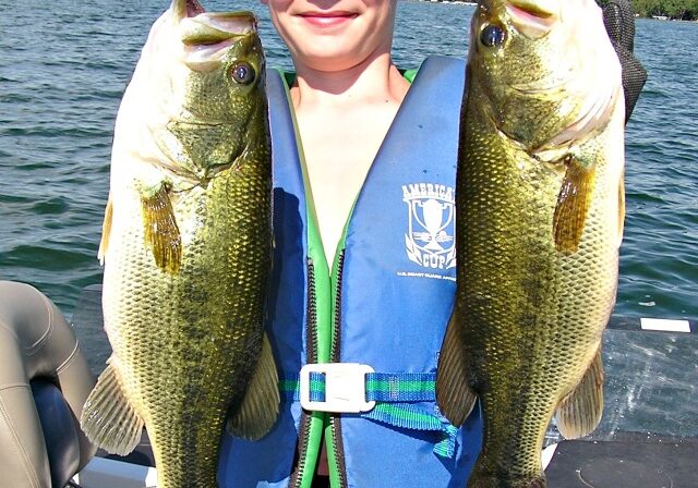 A smiling boy in a life vest and hat stands on a boat, proudly holding up two large fish—proof that Fishing Techniques For Minnesota Anglers really pay off—with a lake and trees visible in the background.