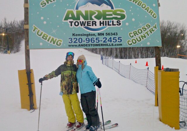 Two people in winter ski gear stand on snow with skis, posing before a "Welcome to Andes Tower Hills" sign at a winter sports area, surrounded by snowy trees and fencing.