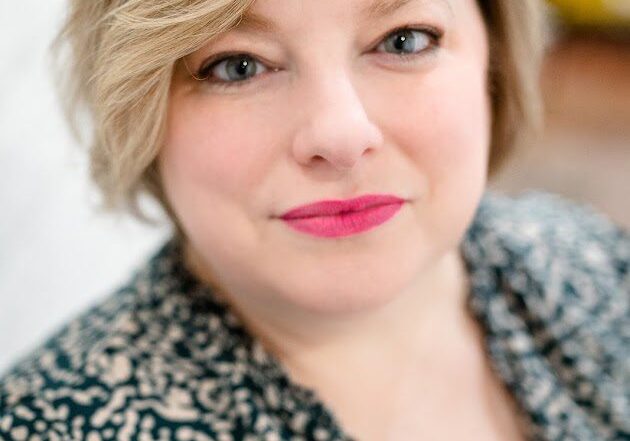 Angela Rantasha, a woman with short, wavy blond hair and pink lipstick, looks at the camera in a patterned black and white top. The softly blurred background reveals hints of white and yellow.