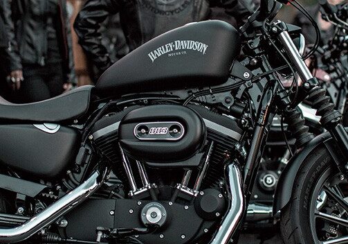 A row of black Apols Harley Davidson motorcycles parked outdoors, with several people nearby dressed in dark clothing. The focus is on the closest motorcycle, highlighting its details and logo clearly.