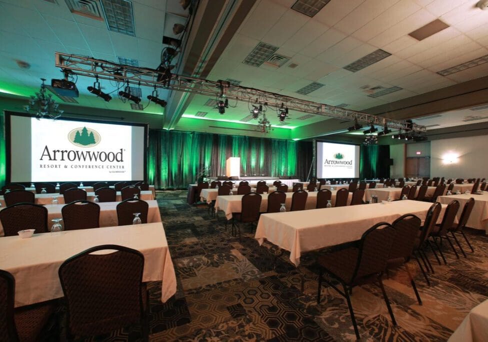 A large conference room at Arrowwood Resort features rows of tables and chairs facing two projection screens displaying the Arrowwood Resort & Conference Center logo. Green lighting highlights the stage and curtains at the front.