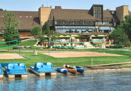A lakeside resort with paddle boats and kayaks by the shore, sandy volleyball courts, green lawns, and a large lodge building with outdoor seating and umbrellas—enjoy all these amenities with Rentals at Arrowwood Resort.