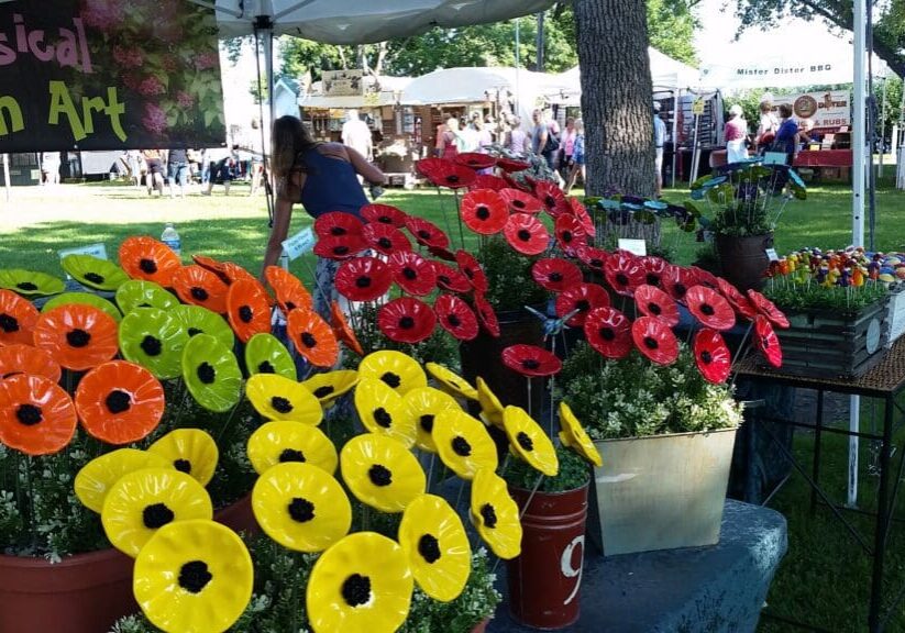 Colorful ceramic flowers in red, orange, and yellow fill pails at an outdoor Art in the Park fair. A sign reads “Whimsical Garden Art” as people wander among tents and booths in the lively background.