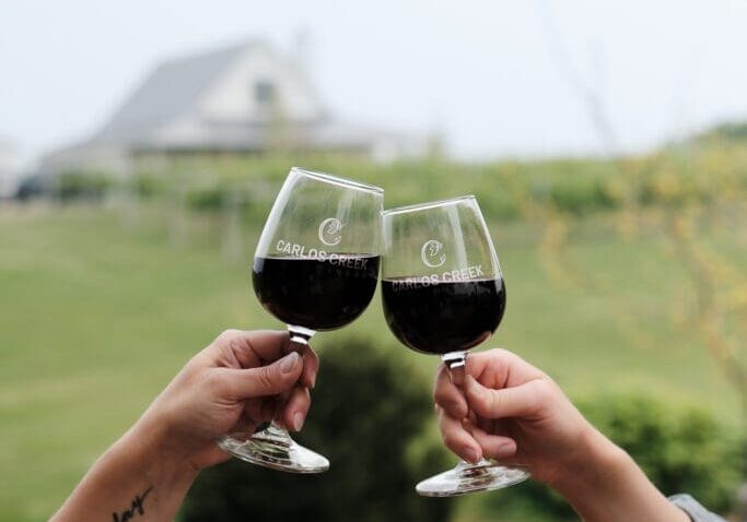 Two people clink glasses of red wine on a wooden deck at Carlos Creek Winery, with a blurred view of a vineyard and house in the background. One person has a tattoo on their forearm.