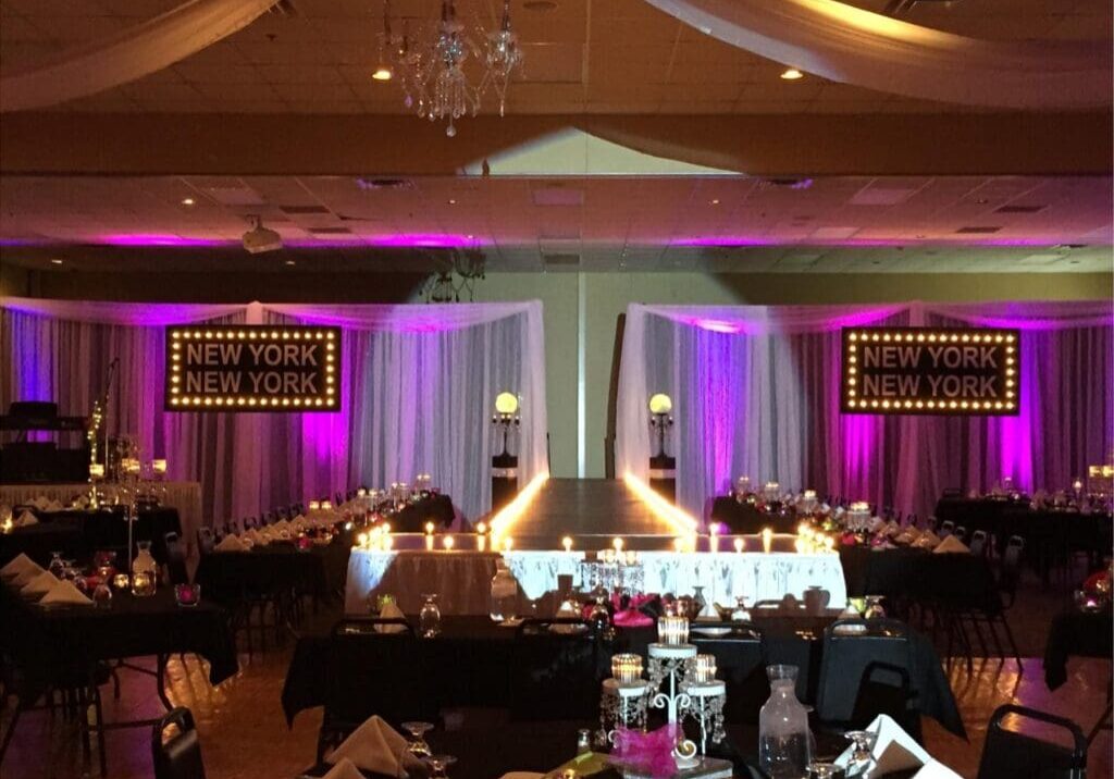 A decorated banquet hall, the Broadway Ballroom, features a lit runway, candle centerpieces, and tables with black tablecloths and napkins. Two large "NEW YORK NEW YORK" signs are displayed on illuminated purple drapes.
