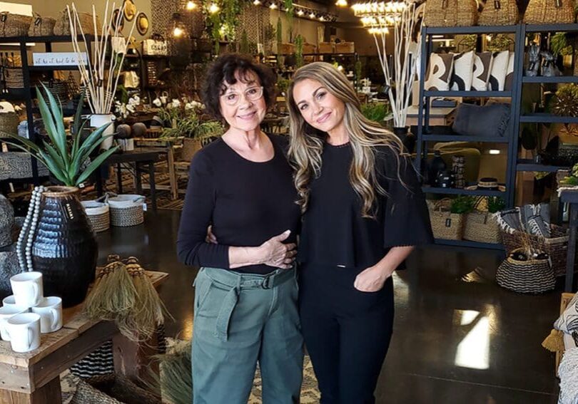 Two women stand smiling together inside Bazara, a stylish downtown Alexandria Minnesota shopping destination filled with plants, baskets, pottery, and decorative items. One woman has short dark hair and glasses; the other has long blonde hair.
