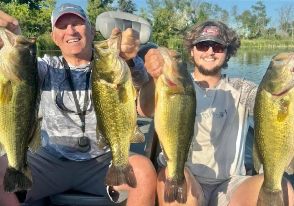 Two people sitting in a boat on a lake, smiling and proudly holding up four large fish they caught after an exciting bass bite, with green trees and blue sky in the background.