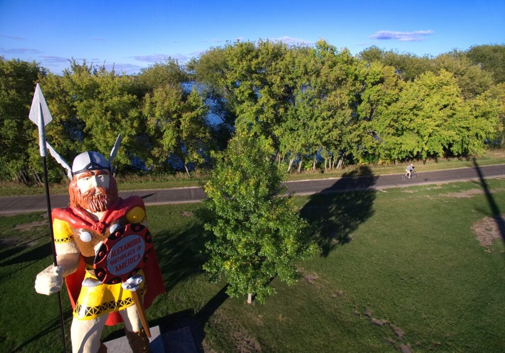 A large Viking statue with a spear and shield stands on grass near a row of trees and a paved path in Big Ole Park Alexandria MN; two people ride bicycles in the background under a clear blue sky.
