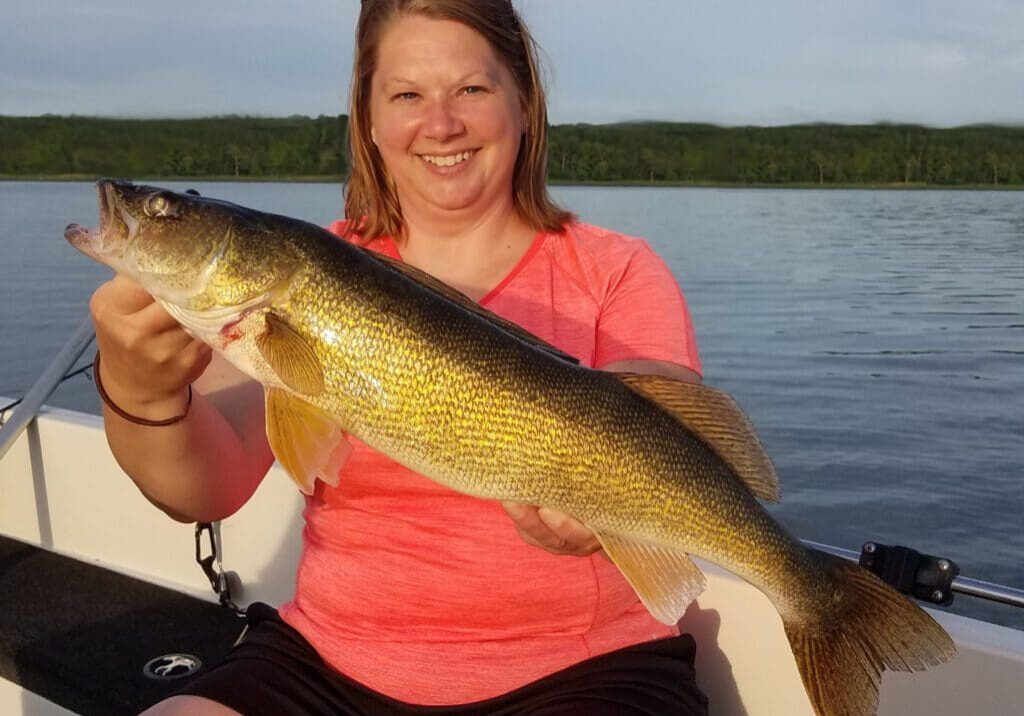 A smiling woman in a bright pink shirt sits on a boat holding a large yellow fish with both hands, with calm water and green trees in the background.
