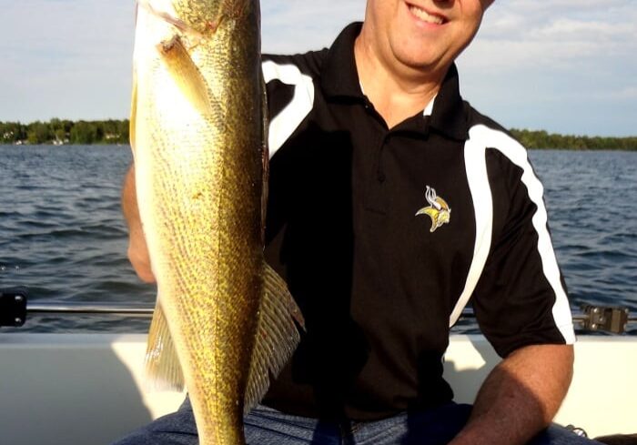Smiling man wearing sunglasses and a cap, sitting in a boat and proudly holding up a large walleye with one hand. Water and distant trees are visible in the background under a blue sky.