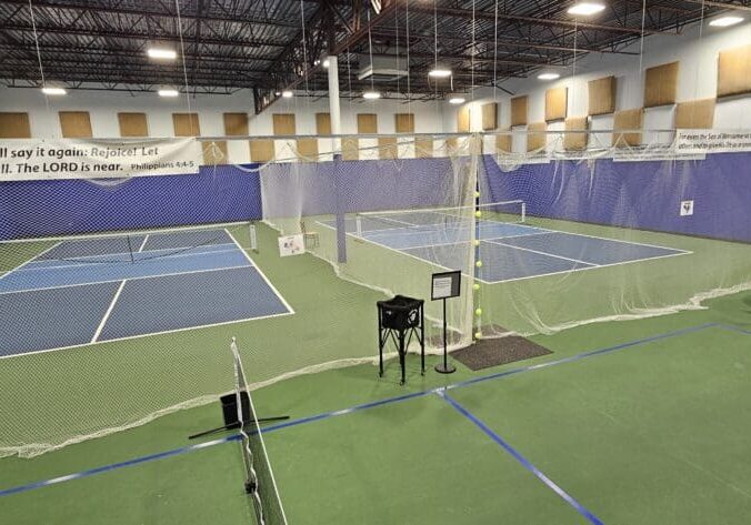Indoor sports facility featuring multiple pickleball courts at Blind Squirrel Pickleball Club, with green flooring, netting between courts, tables and chairs on the right, and a biblical banner hanging above the courts.